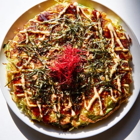 A close-up of a fluffy Japanese Okonomiyaki Pancake garnished with green onions and pickled ginger on a rustic wooden plate.