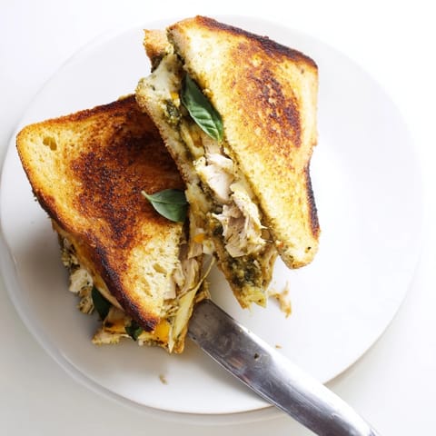 Sliced Mozzarella Pesto Chicken Grilled Cheese on a rustic board, revealing layers of shredded chicken, vibrant green pesto, and fresh tomato.